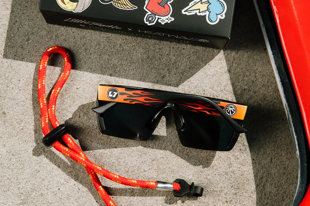 Backside shot of the Kids Lazer Face Sunglasses: Little Trouble X Heat Wave Flames with strap and box on a concrete floor.