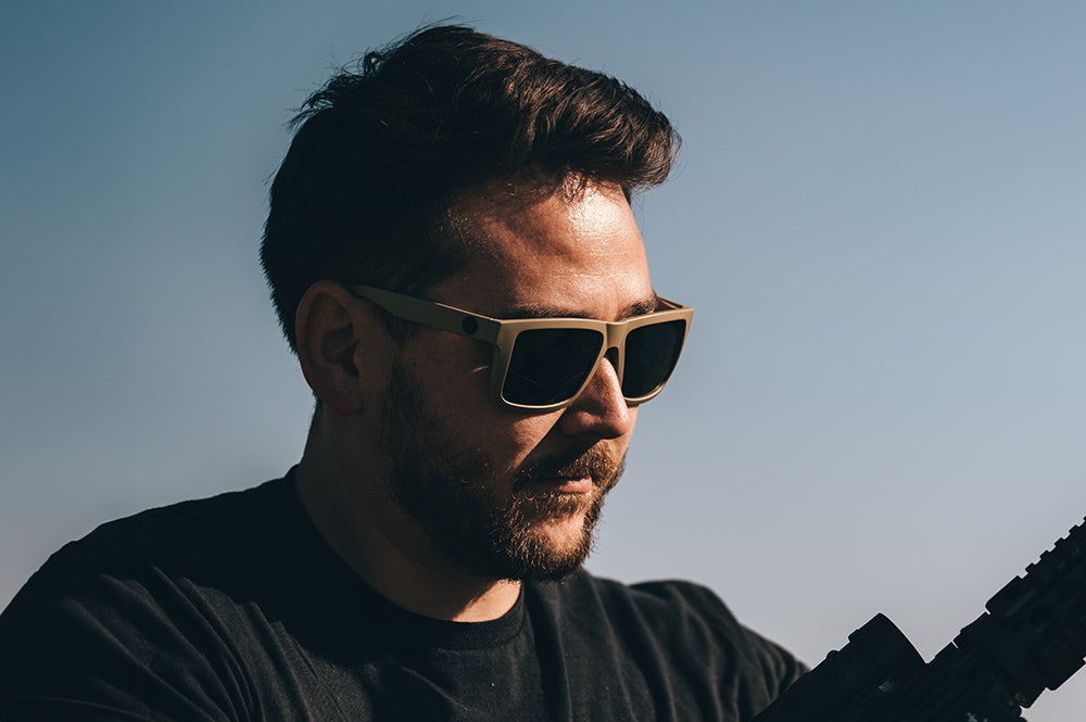 Man wearing Heat Wave XL Vise tan sunglasses with ultra black lenses and a black shirt holding a rifle against a clear sky.