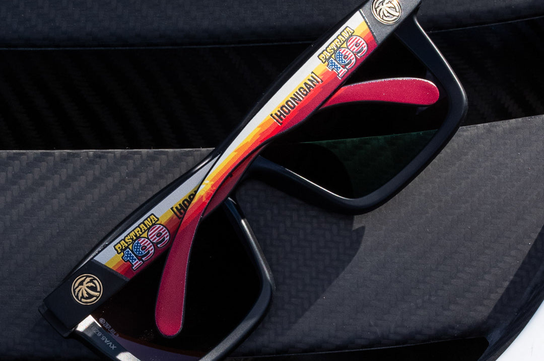 Lying on carbon fiber is the Heat Wave Visual XL Vise Hoonigan Pastrana Sunglasses. 