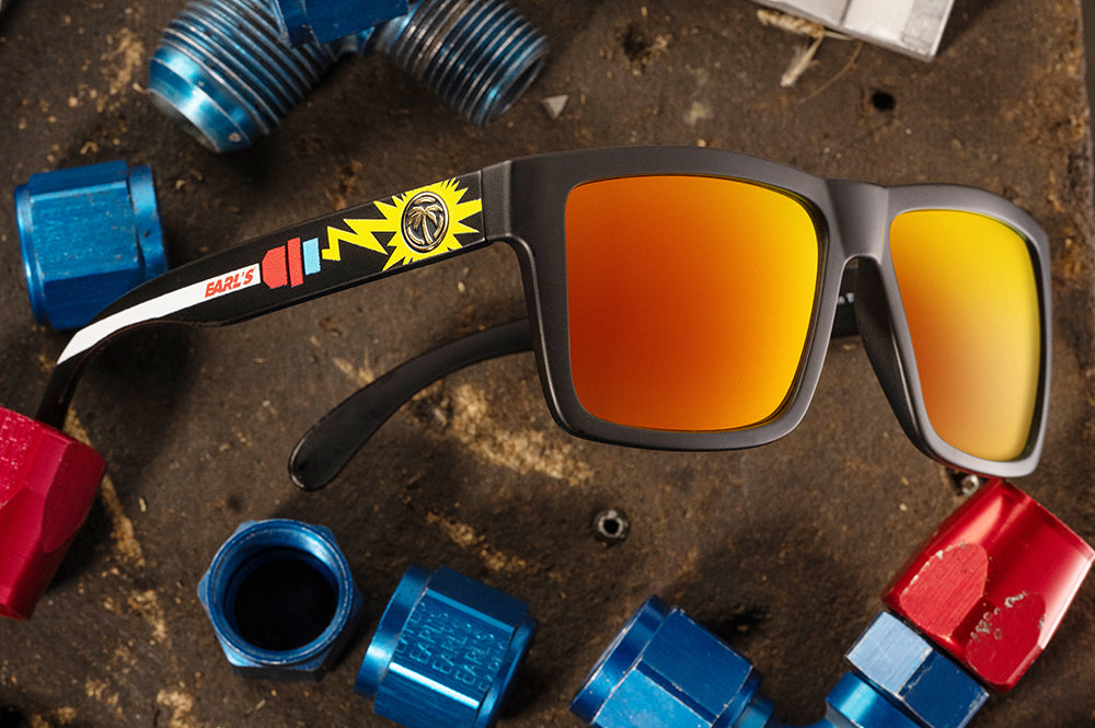 Lying on the ground is the Heat Wave Visual Vise Sunglasses with black frame, Earl's x HWV collab and sunblast orange yellow lenses.